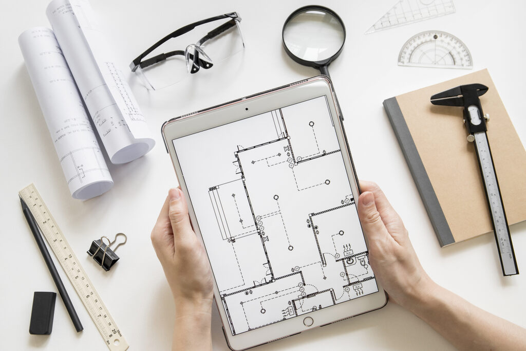 Plano técnico digital de instalación eléctrica mostrado en una tablet sostenida por dos manos, rodeada de herramientas de arquitectura como planos enrollados, regla, transportador, calibrador, cuaderno y lupa.