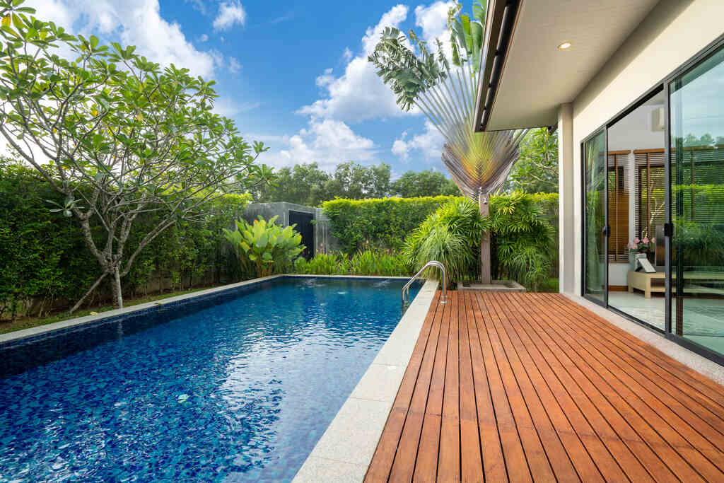 Linda piscina rodeada de plantas verdes e árvores, com um deck de madeira, em um dia ensolarado, perfeito para relaxar e curtir o verão.