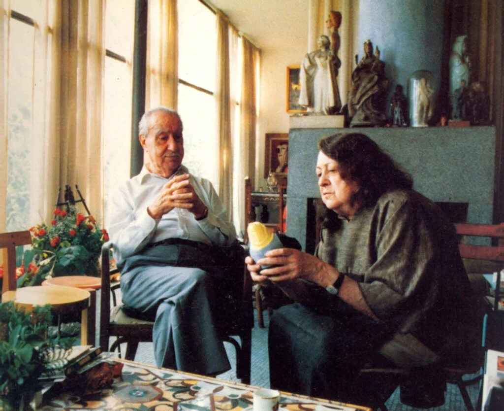 Fotografía en color de Lina Bo Bardi y su esposo Pietro Maria Bardi en el interior de la Casa de Vidrio en São Paulo. La pareja está sentada en un acogedor espacio lleno de arte; Lina observa un objeto mientras Pietro sostiene una taza, rodeados de elementos decorativos y la luz natural que entra por los grandes ventanales.