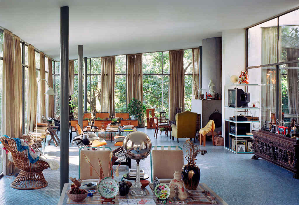 Vista del interior de la Casa de Vidrio en São Paulo, diseñada por Lina Bo Bardi. La amplia sala de estar cuenta con ventanales de suelo a techo que conectan el espacio con la vegetación exterior, llena de muebles eclécticos, objetos de arte y piezas decorativas que reflejan el estilo personal y artístico del matrimonio Bardi.