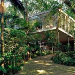 View of the iconic Glass House in São Paulo, Brazil, surrounded by lush tropical vegetation. The modernist residence, designed by architect Lina Bo Bardi, features floor-to-ceiling glass walls elevated on slender stilts above the forested landscape.
