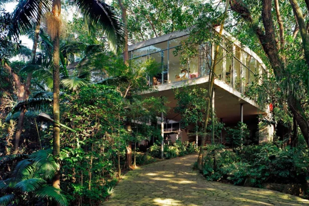 View of the iconic Glass House in São Paulo, Brazil, surrounded by lush tropical vegetation. The modernist residence, designed by architect Lina Bo Bardi, features floor-to-ceiling glass walls elevated on slender stilts above the forested landscape.
