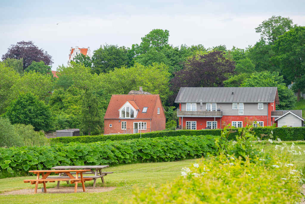 Rural landscaping with two country-style houses surrounded by lush greenery, dense trees, hedges, and a picnic table on a grassy area in the foreground.