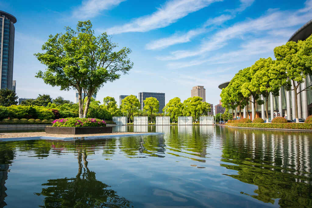 Urban landscaping with a large reflective water feature, lush trees, flower beds, and modern buildings in the background under a blue sky with light clouds.