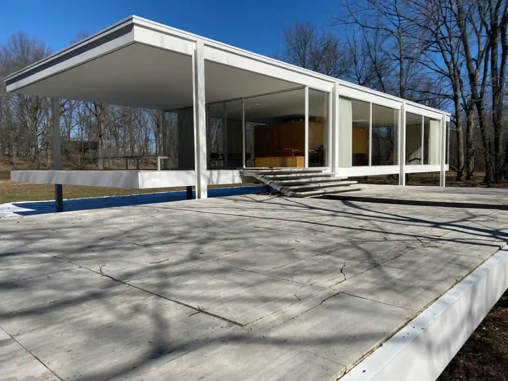 Imagem da casa Farnsworth, uma residência moderna com estilo minimalista, estrutura de vidro e concreto, localizada em ambiente ao ar livre com árvores ao fundo.