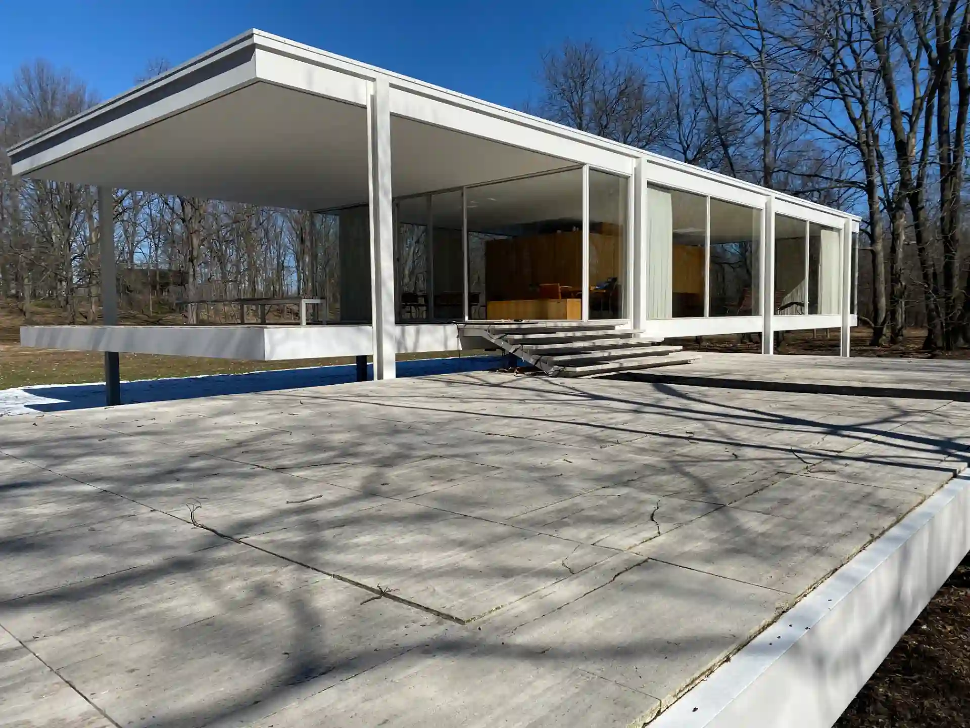 Imagem da casa Farnsworth, uma residência moderna com estilo minimalista, estrutura de vidro e concreto, localizada em ambiente ao ar livre com árvores ao fundo.