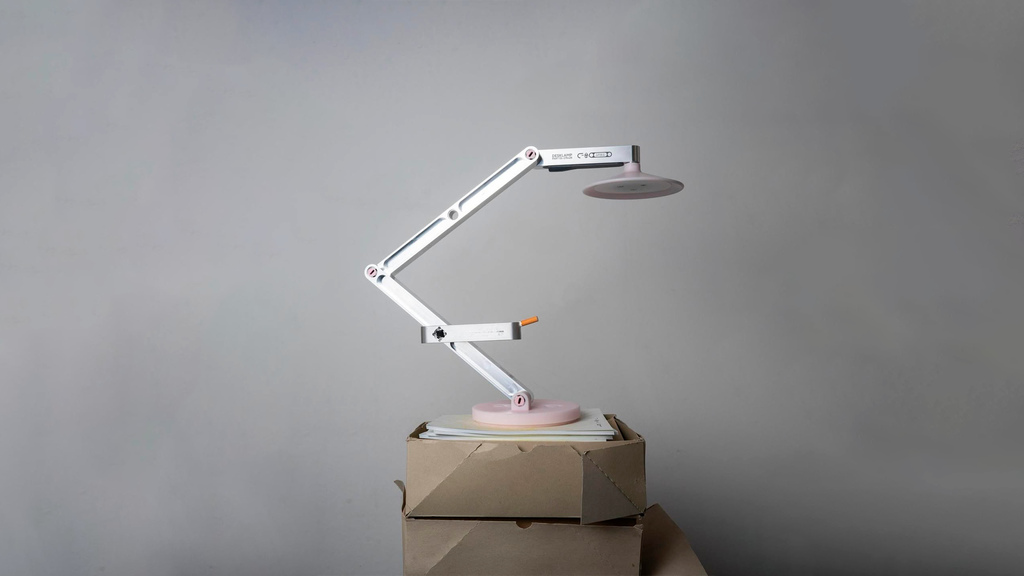 Luminária de mesa articulada de design moderno, branca com detalhes em rosa, sobre caixas, refletindo tendências para o Salão de Milão 2026.