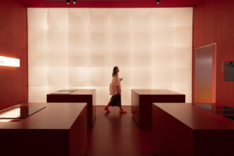 Mulher em estande moderno com balcões minimalistas vermelhos e parede iluminada, representando tendências de design de interiores e inovação tecnológica esperadas no Salão de Milão 2026.