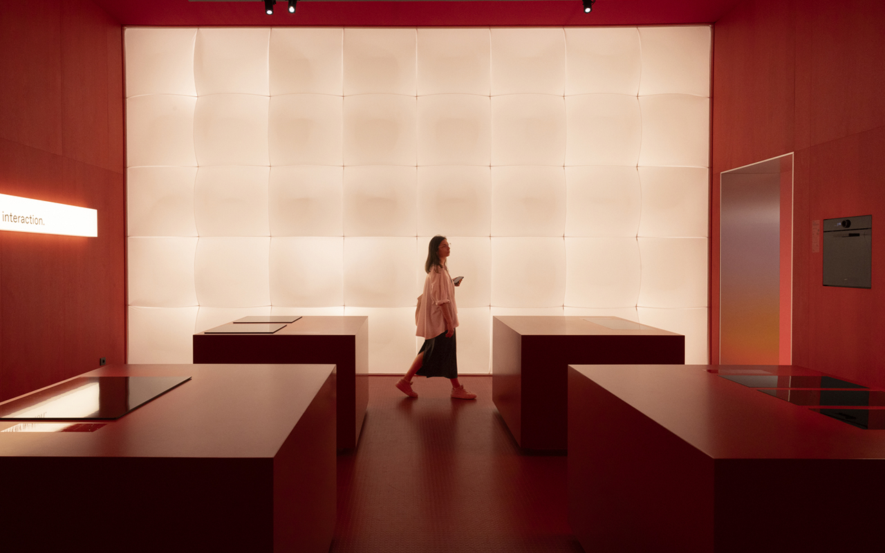 Mulher em estande moderno com balcões minimalistas vermelhos e parede iluminada, representando tendências de design de interiores e inovação tecnológica esperadas no Salão de Milão 2026.