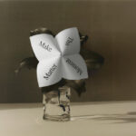 Dobradinha de papel com as palavras "Make This Moment Matter" em vaso de vidro com folhas escultóricas escuras, fotografada em tons quentes e sombrios — imagem tema do 3 Days of Design 2026.