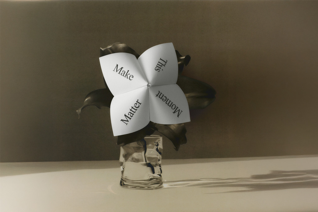 Dobradinha de papel com as palavras "Make This Moment Matter" em vaso de vidro com folhas escultóricas escuras, fotografada em tons quentes e sombrios — imagem tema do 3 Days of Design 2026.