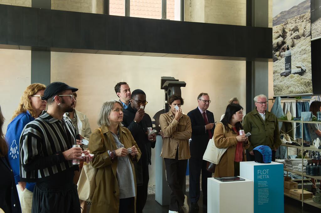 Grupo de visitantes reunidos em espaço expositivo durante o 3 Days of Design 2026, com bebidas nas mãos e atenção voltada para apresentação. Ao fundo, placa de exposição da marca Flot Hetta, prateleiras com produtos e imagem fotográfica de paisagem natural.
