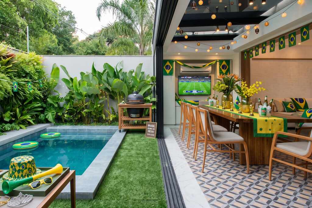 Área externa com piscina decorada para a copa do mundo 2026, com bandeirinhas do Brasil, luzinhas amarelas e mesa em verde e amarelo.