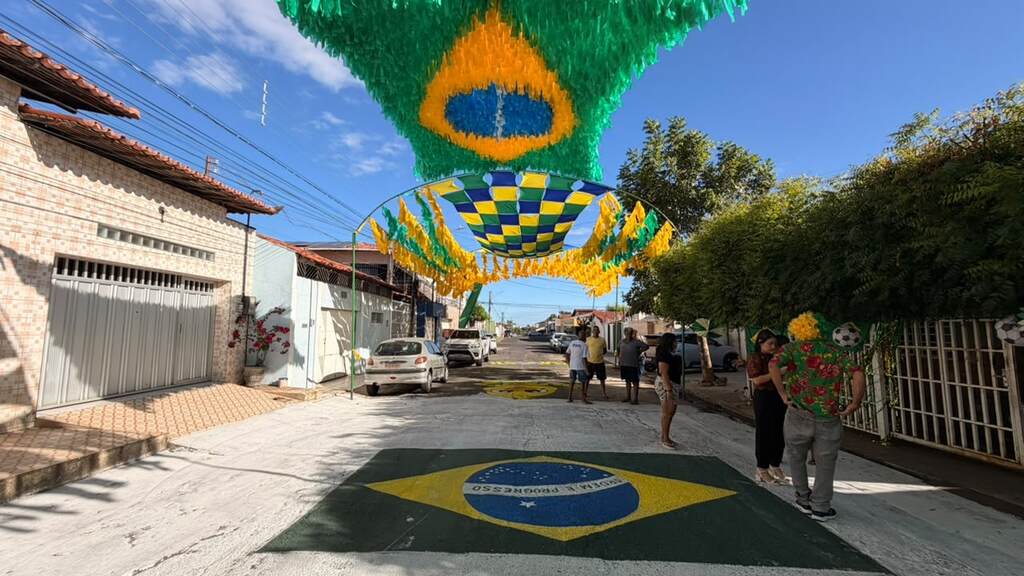 Rua brasileira vibrantemente decorada para a Copa do Mundo 2026, com a bandeira do Brasil pintada no asfalto e grandes ornamentos aéreos em verde, amarelo e azul, criando um cenário festivo.