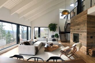 Ampla sala de estar com design de interiores em estilo escandinavo, destacando-se pela abundante luz natural, teto com vigas brancas, sofá modular claro, lareira moderna com revestimento de madeira rústica e vista panorâmica para a floresta.