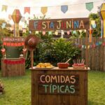 Ambiente externo festivo com diversas ideias de decoração para festa junina, apresentando bandeirinhas coloridas, balões juninos, barracas de madeira com comidas típicas, jogos como acerte a lata e fogueira.