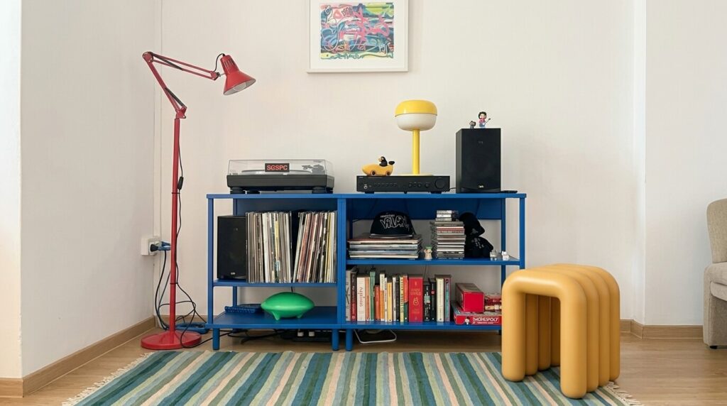 Ambiente de sala de estar vibrante e personalizado com estante azul repleta de discos de vinil, toca-discos, livros e objetos decorativos ecléticos, incluindo uma luminária amarela e um pufe mostarda, refletindo o descontraído e divertido estilo Funhaus.