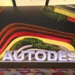 Letreiro luminoso AUTODESK em destaque na entrada de um evento, com um fundo de parede vibrante de design orgânico e elementos verdes, capturando o ambiente da Autodesk University 2026.