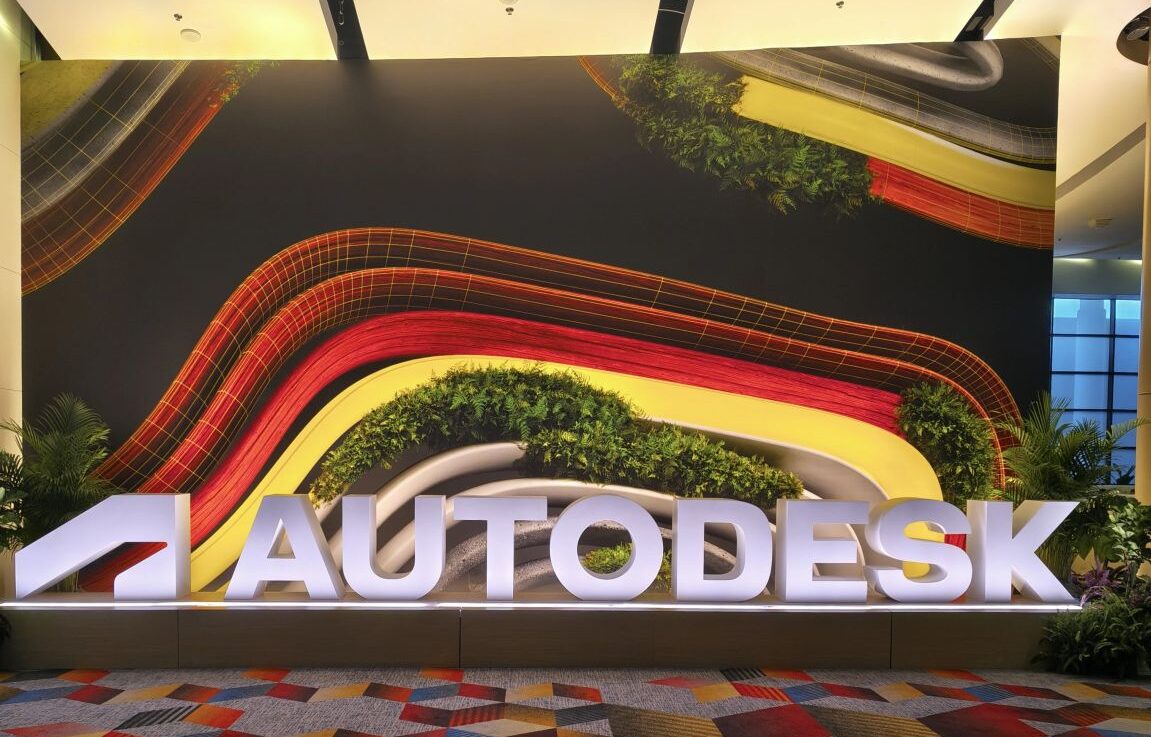 Letreiro luminoso AUTODESK em destaque na entrada de um evento, com um fundo de parede vibrante de design orgânico e elementos verdes, capturando o ambiente da Autodesk University 2026.